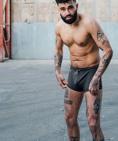 Man with tattoos wearing black swim shorts standing on a concrete surface.